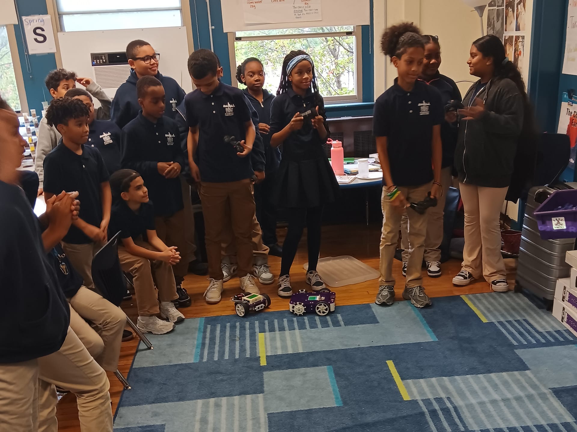 A class of students stands in a circle on the classroom carpet with game controllers while two robot kits drive between them
