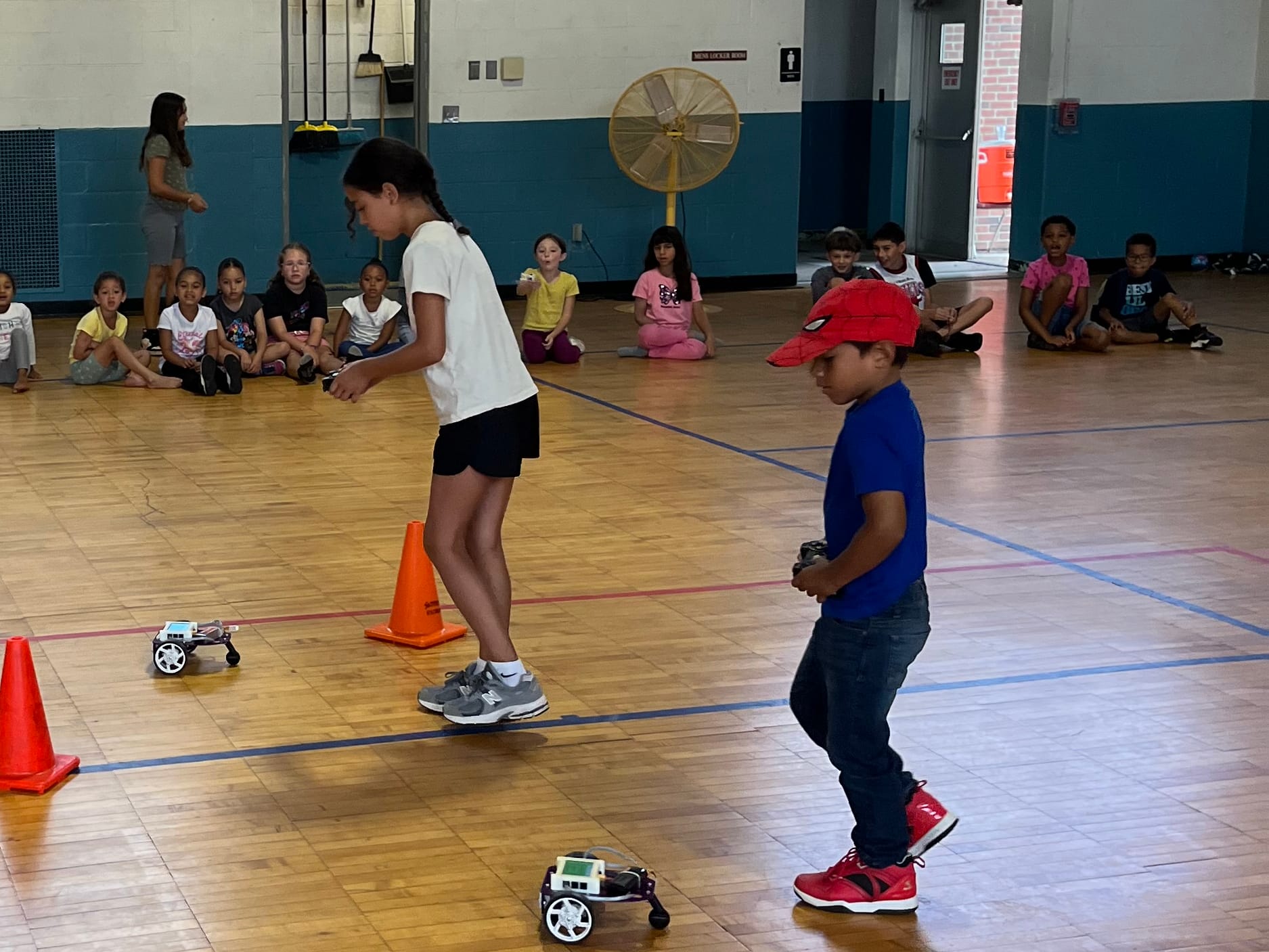 Two young students drive Pilot robots around orange cones on a gym floor while campers watch from the sideline