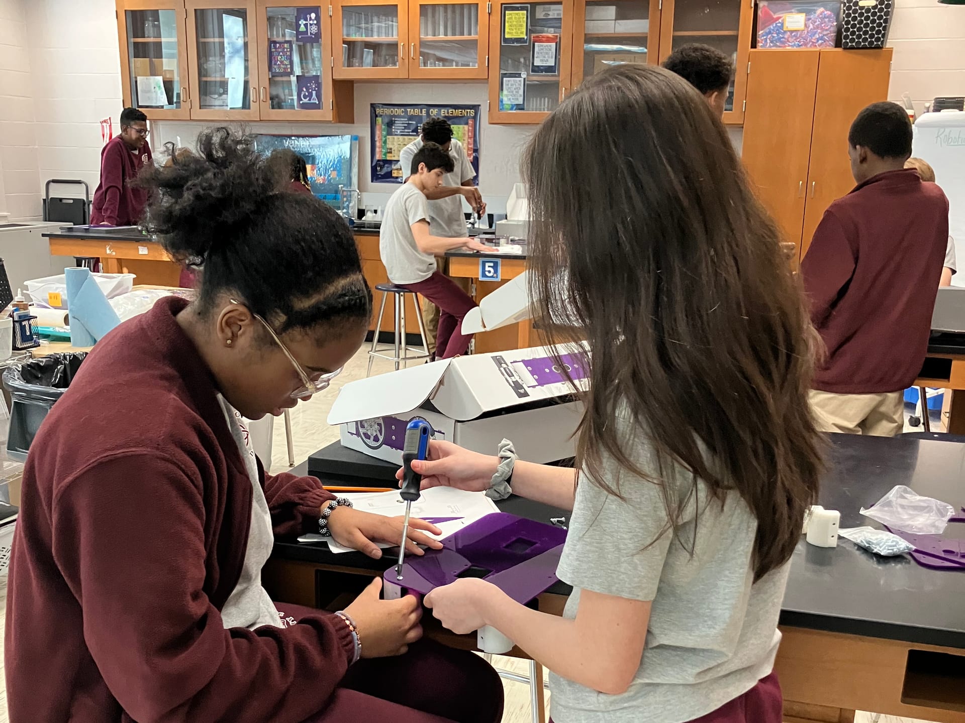 Two middle school students assemble a purple Axle Robot Kit chassis with a screwdriver at a science lab bench